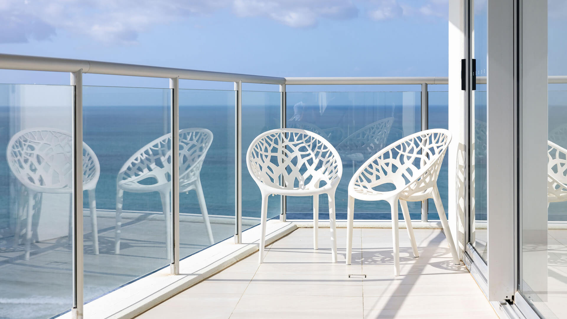 Balcony with white chairs and glass railing overlooking the ocean in ULTIQA Air’s 3 Bedroom Ocean View Suite.