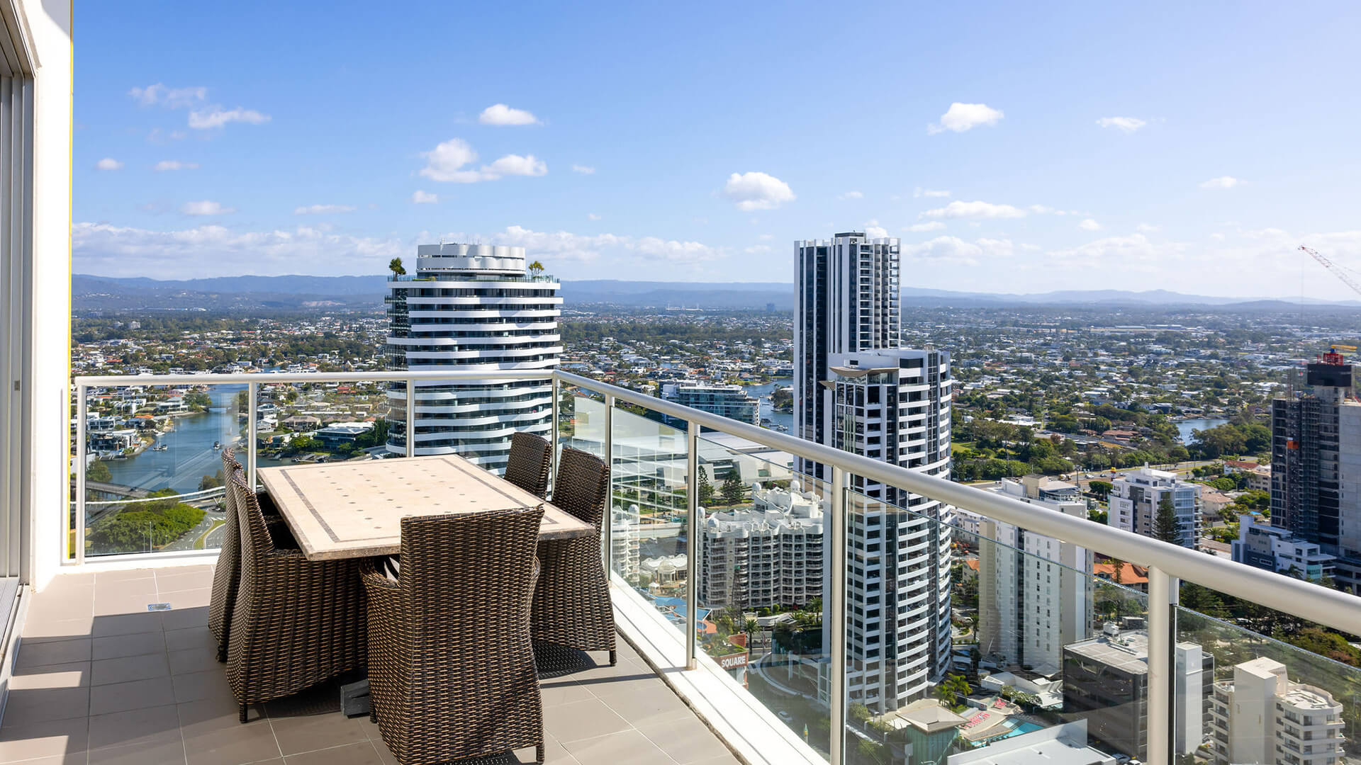 High-rise balcony with wicker dining set and glass railing overlooking Gold Coast cityscape and river.