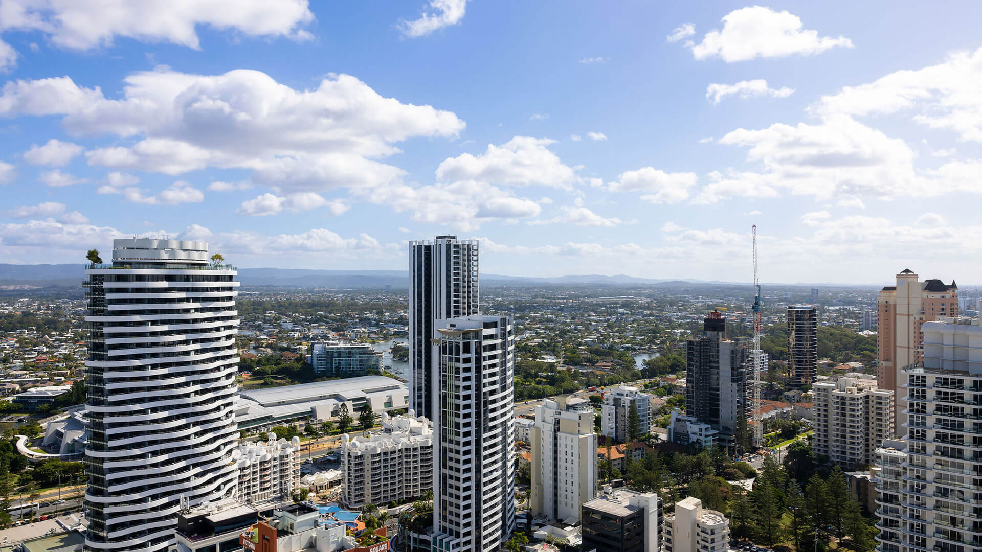 Cityscape with high-rise buildings, river, and distant mountains under a partly cloudy sky.