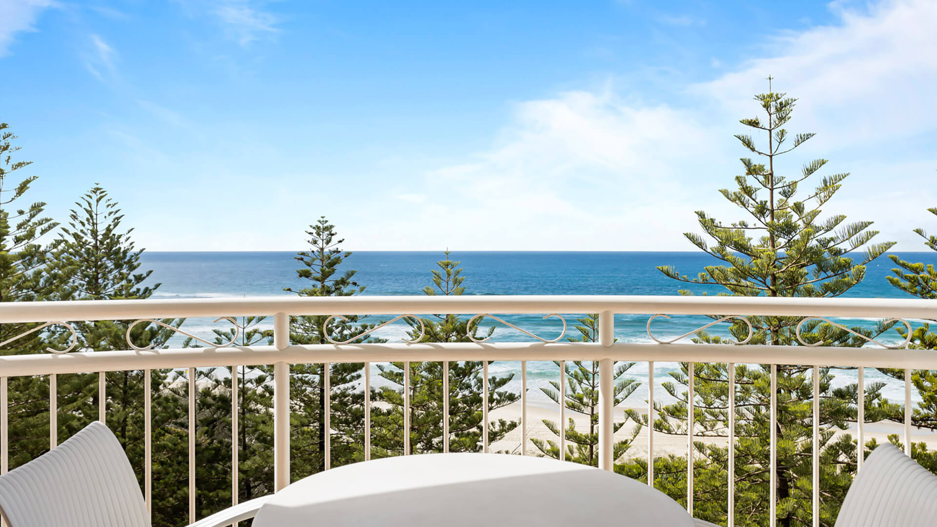 Ocean-view balcony with outdoor seating overlooking the coastline, greenery, and surrounding buildings.