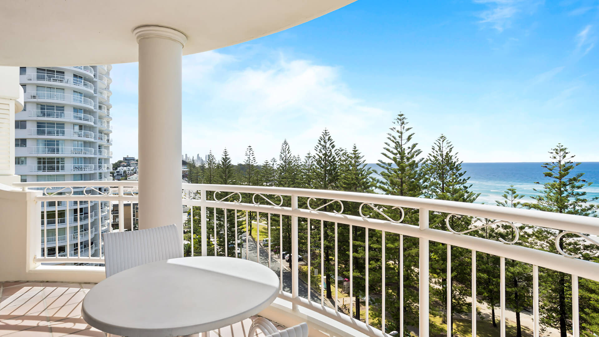 Ocean-view balcony with outdoor seating overlooking the coastline, greenery, and surrounding buildings.